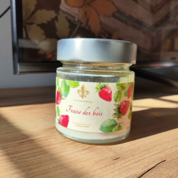 Bougie artisanale Fraise des bois Les Lueurs d’Horion en verre avec couvercle en métal, posée sur une surface en bois, éclairée par le soleil.