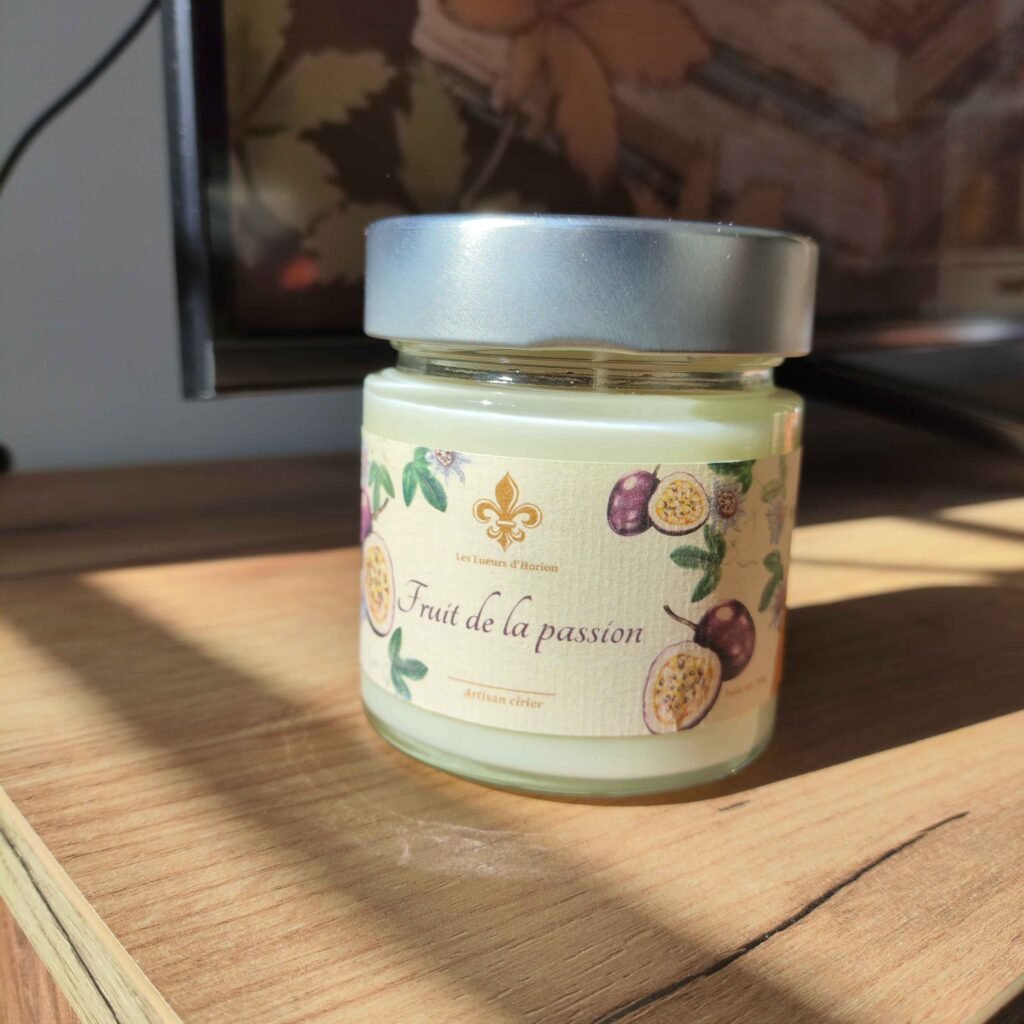 Bougie artisanale Fruit de la passion Les Lueurs d’Horion en verre avec couvercle en métal, posée sur une surface en bois, éclairée par le soleil.