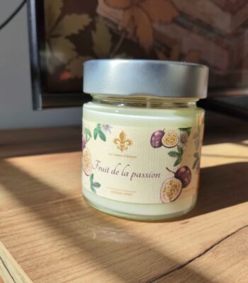 Bougie artisanale Fruit de la passion Les Lueurs d’Horion en verre avec couvercle en métal, posée sur une surface en bois, éclairée par le soleil.