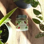 Bougie artisanale Fraise des bois Les Lueurs d’Horion posée sur un sol en bois, entourée de plantes vertes, éclairée par la lumière naturelle.