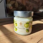 Bougie artisanale Pomme verte Les Lueurs d’Horion en verre avec couvercle en métal, posée sur une surface en bois, éclairée par le soleil.